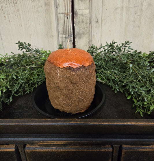 A vintage orange and clove scented hearth candle placed on a black dish, with a rustic wooden background and greenery around it.
