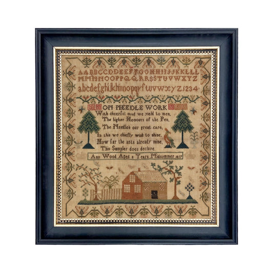 A framed needlepoint sampler featuring the alphabet, numbers, and a historical house embroidery, with trees and birds depicted.