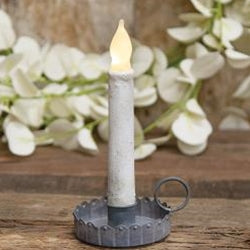 A taper candle in an antiqued gray scalloped taper holder with a fluted design and a round loop handle, placed on a wooden surface with white flowers in the background.