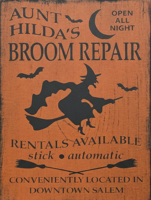 Wooden sign with witch silhouette and text about broom repair, surrounded by greenery.