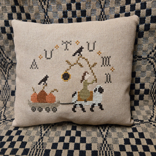 A square beige woven throw pillow featuring a cross-stitched autumn-themed design with a sheep, cart, and pumpkins displayed on a black and white checkered patterned couch.
