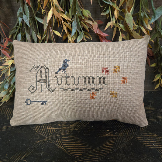 Decorative pillow with 'Autumn' stitching on a textured surface with leaves.