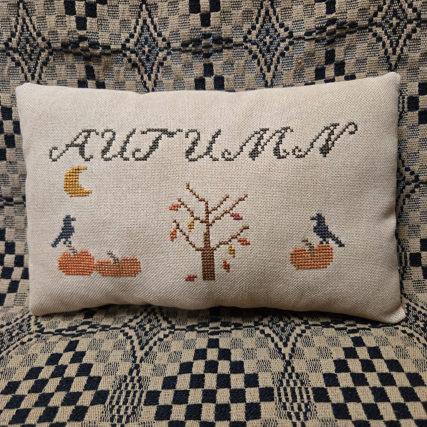 A square beige linen throw pillow with a cross-stitched autumn design featuring the word 'AUTUMN', a tree, pumpkins, crows, and a moon, resting on a patterned blue and beige couch.
