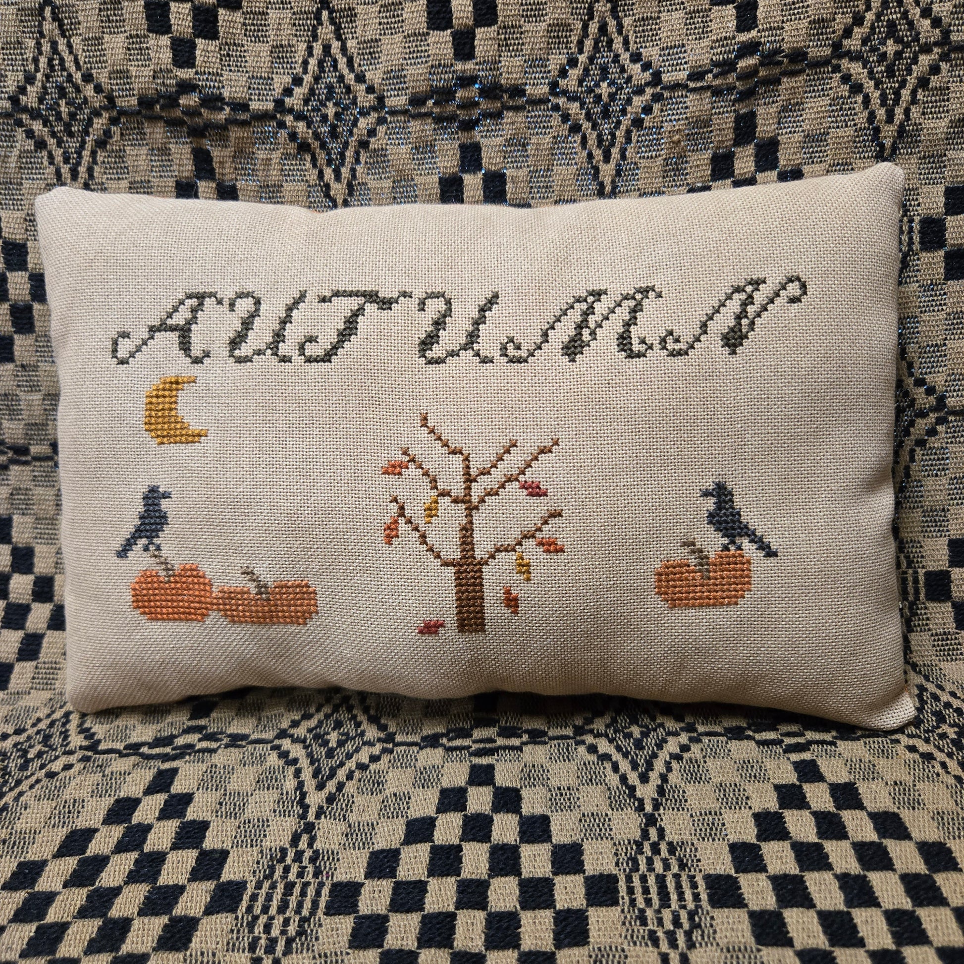 A square beige linen throw pillow with a cross-stitched autumn design featuring the word 'AUTUMN', a tree, pumpkins, crows, and a moon, resting on a patterned blue and beige couch.
