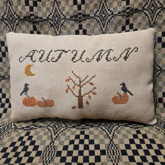 A square beige linen throw pillow with a cross-stitched autumn design featuring the word 'AUTUMN', a tree, pumpkins, crows, and a moon, resting on a patterned blue and beige couch.
