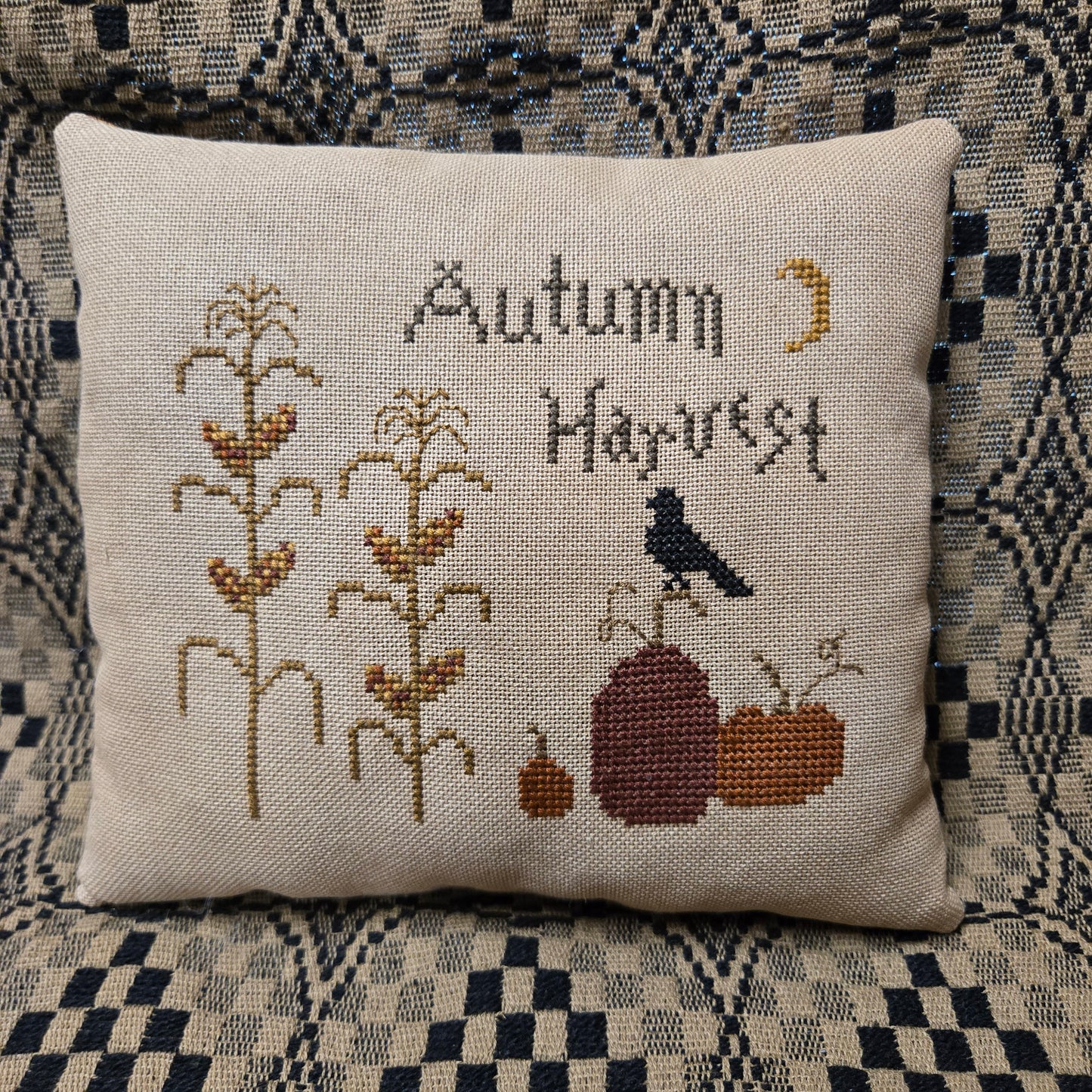 A square linen throw pillow featuring a cross-stitch design with the words 'Autumn Harvest,' corn stalks, pumpkins, a bird, and a crescent moon, placed on a patterned blue and beige couch.