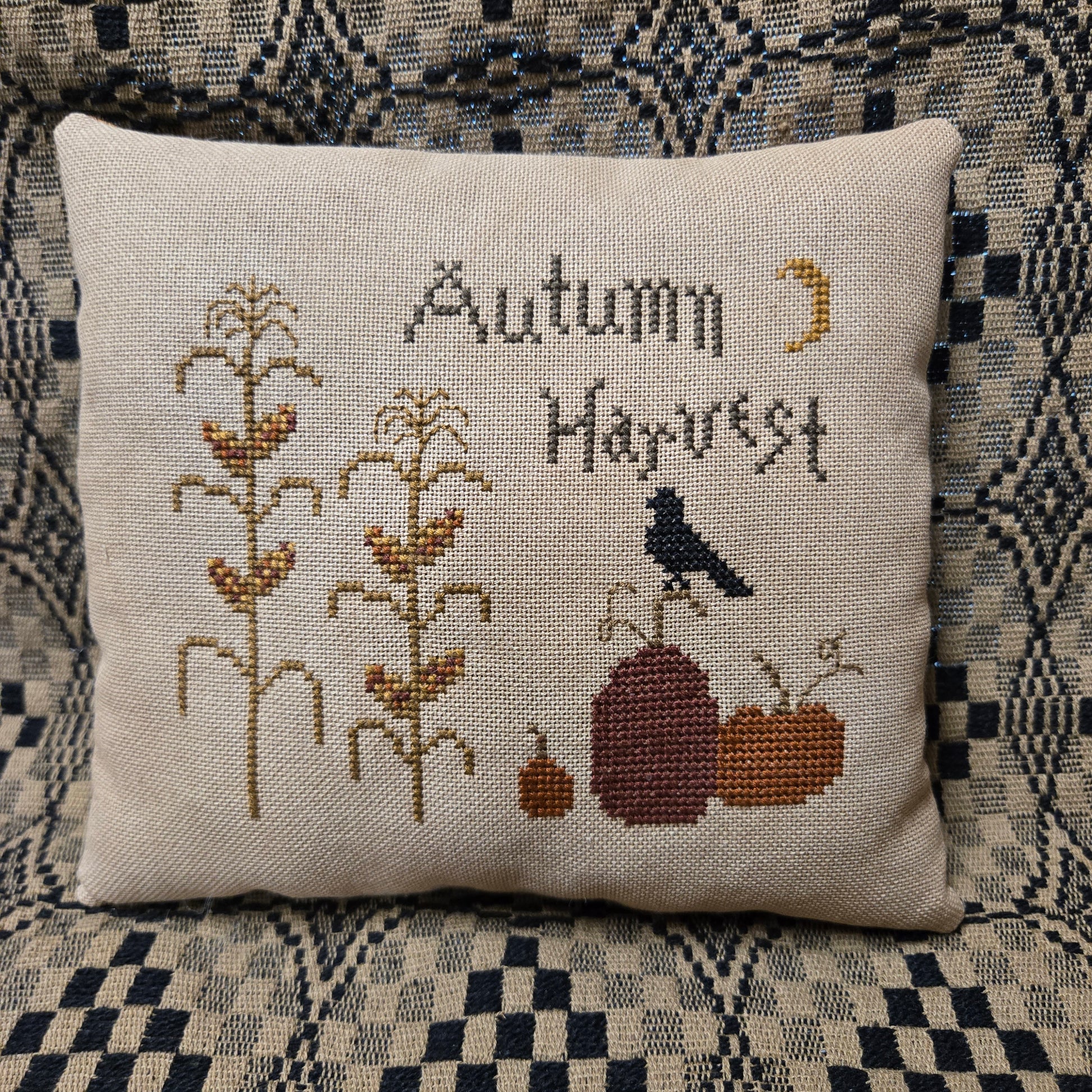 A square linen throw pillow featuring a cross-stitch design with the words 'Autumn Harvest,' corn stalks, pumpkins, a bird, and a crescent moon, placed on a patterned blue and beige couch.