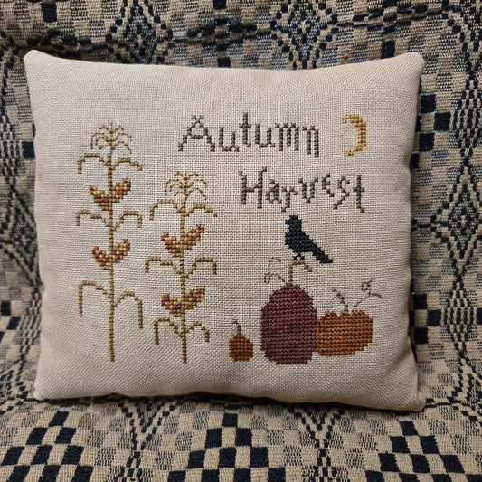 A square linen throw pillow featuring a cross-stitch design with the words 'Autumn Harvest,' corn stalks, pumpkins, a bird, and a crescent moon, placed on a patterned blue and beige couch.