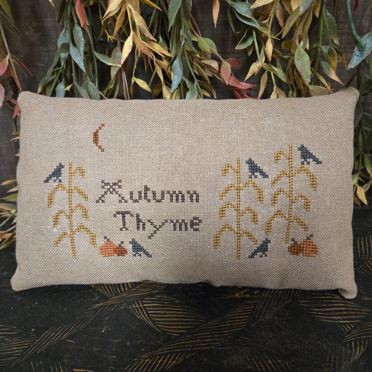 Cross-stitched pillow with 'Autumn Thyme' text and floral patterns on a textured surface.