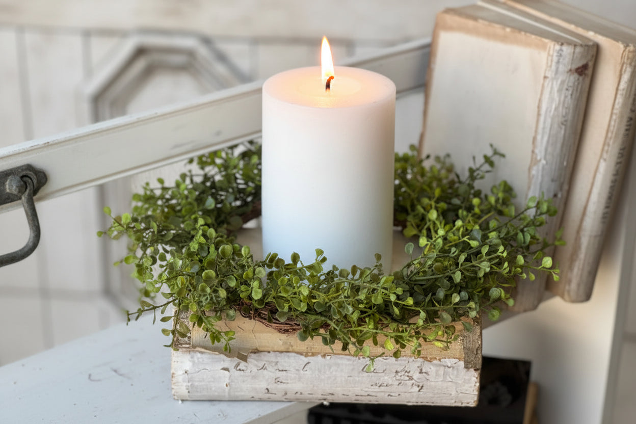 White candle on a wooden block with greenery, placed on a surface.