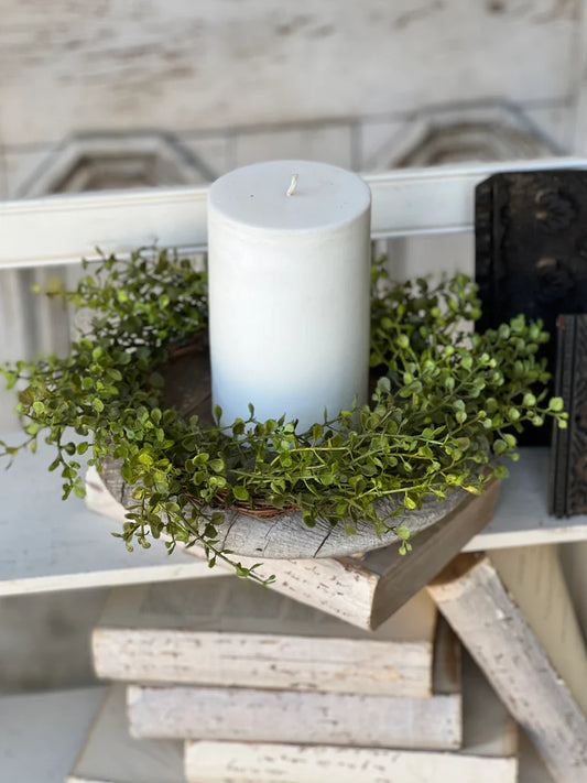 A candle placed in the center of a green candle ring, which is resting on a wooden piece. Around the ring, there are small round green leaves.