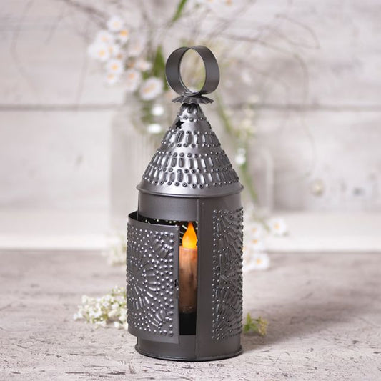 A smokey black traditional round lantern with punch designs and a lit candle inside, placed on a white surface with a floral arrangement in the background.