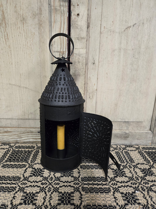 A traditional round black lantern with punch designs and a textured finish, displaying a lit candle inside, placed on a patterned textile surface.