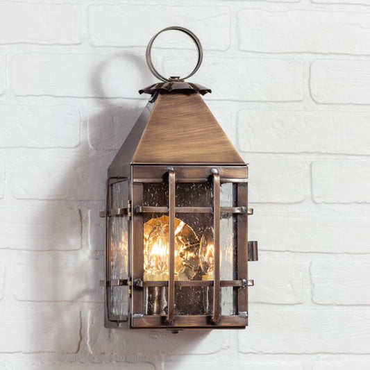 A solid weathered brass outdoor wall light with a lantern design, featuring clear glass panels and an metallic exterior, mounted on a white brick wall.