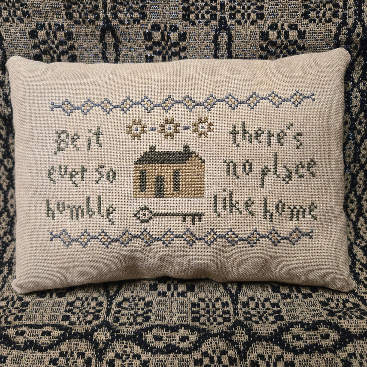 Decorative pillow with cross-stitched text and a house design on a patterned background