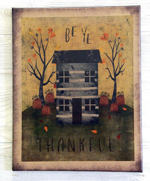 A rustic framed art print featuring a dark gray log cabin, orange pumpkins, bare trees with orange leaves, and the words 'BE YE THANKFUL' on a weathered yellow and brown background.