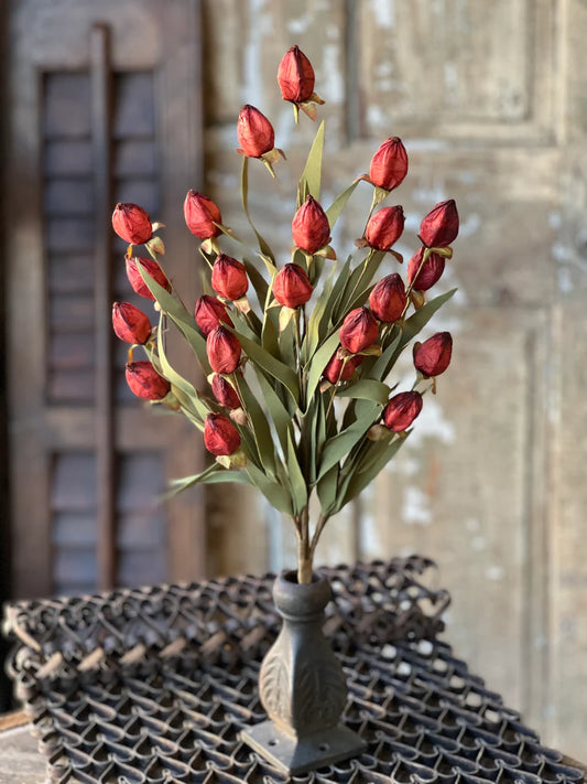 A faux floral arrangement resembling a bouquet of lantern-like red buds with greenery, placed in a gray vase, set against a rustic backdrop.