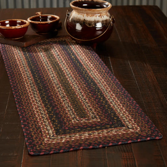 A rectangular braided table runner with concentric rectangular patterns in brown, red, black, and beige tones, displayed on a dark wooden table with ceramic bowls and a pot in the background.
