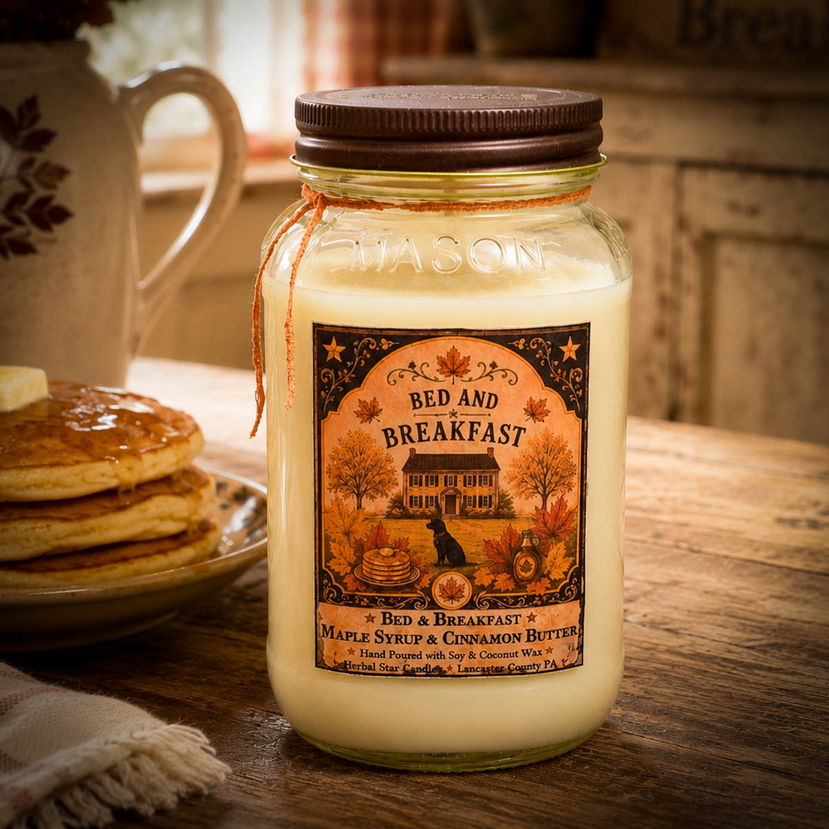 Jar of 'Bed and Breakfast' candle with a label on a wooden table with pancakes and a teapot.
