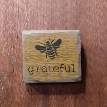 A rectangular wooden sign with a bee illustration and the word 'grateful' inscribed on it, placed on a wooden surface.