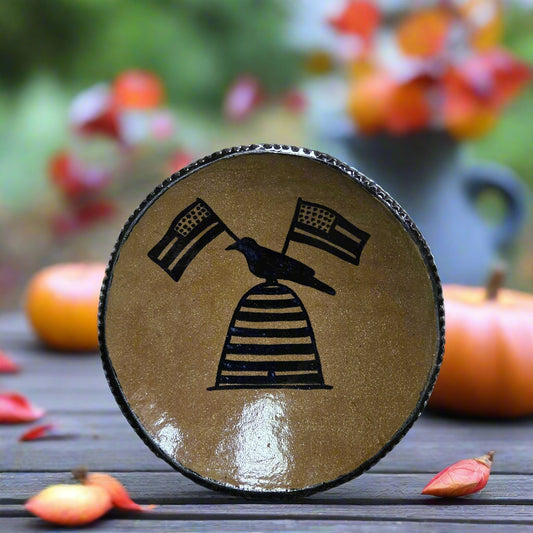 Decorative wooden slice with a bird and flag design on a wooden surface with pumpkins and flowers in the background.