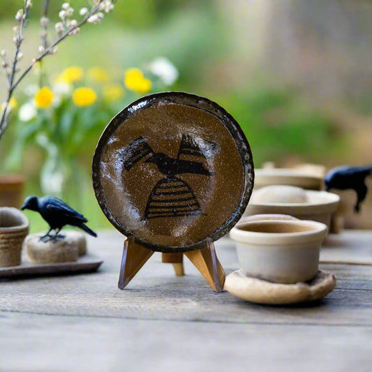 Ceramic dish with a bird design on a wooden stand, surrounded by other ceramic items on a blurred natural background.