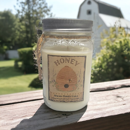 A 16 oz mason jar labeled 'HONEY' filled with a candle, set on a wooden surface with a farmhouse in the background.