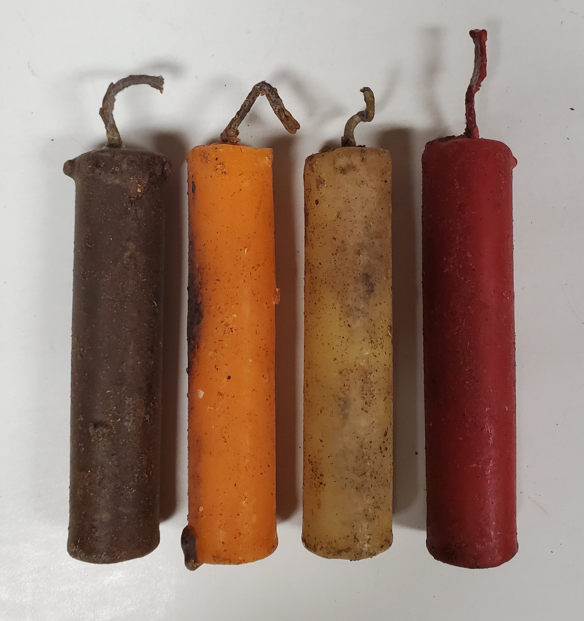 A set of four beeswax candle tapers in brown, pumpkin, ivory, and red colors, each with a primitive-looking wick and end cap.