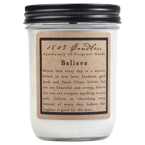 A white jar candle with a label that reads 'Believe' and has a quote underneath. The candle is placed in a clear jar with a black lid.