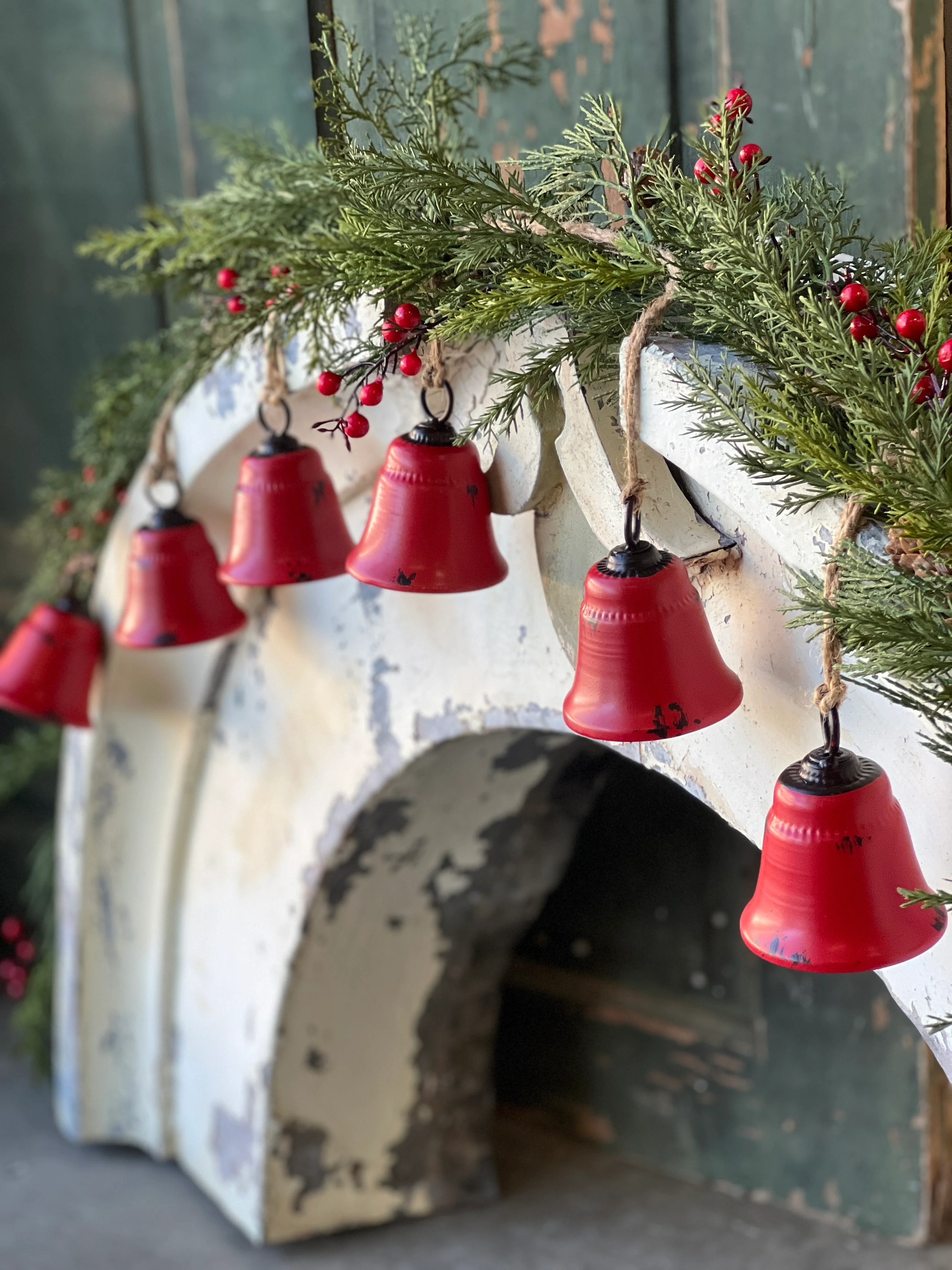 A garland of red metal bells hanging from a white structure, with greenery and red berries adding to the decor.