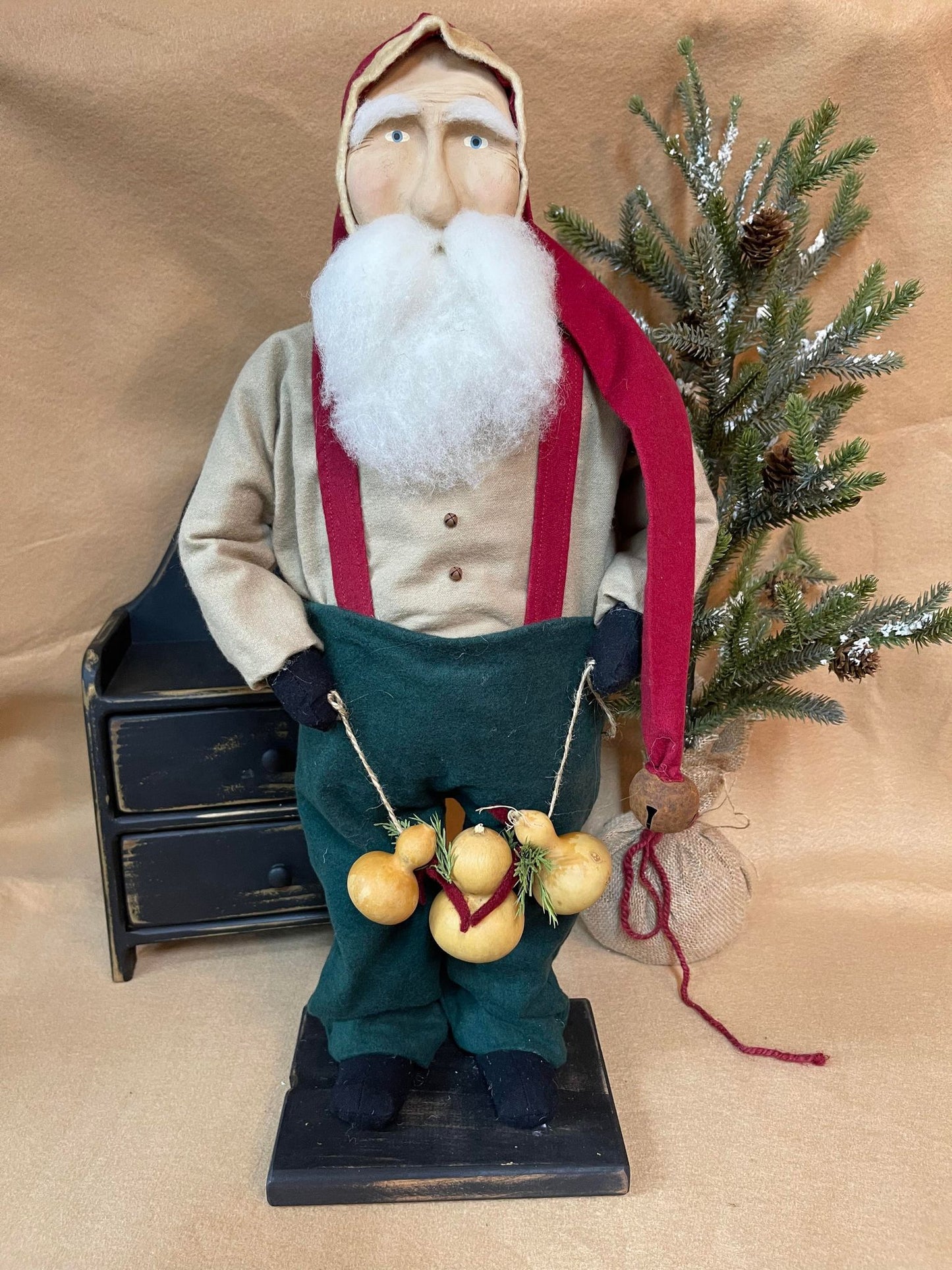 A Belsnickel figurine holding a gourd garland, with a Santa hat and coat, standing next to a decorative tree.