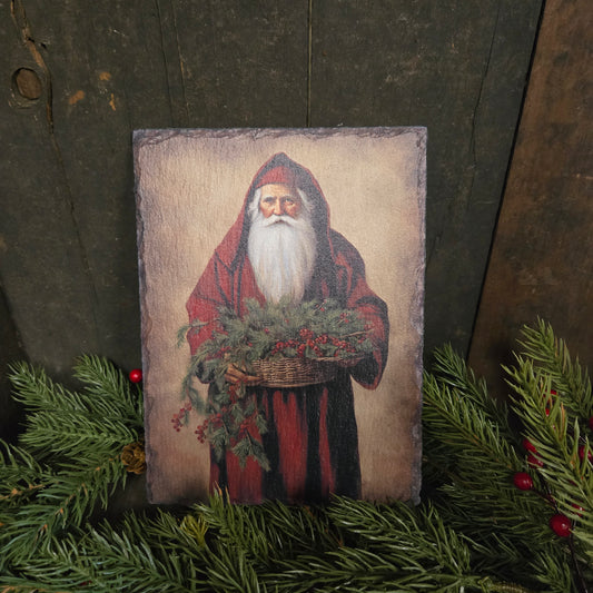 Slate plaque with a Santa Claus illustration on a rustic wooden surface with greenery.
