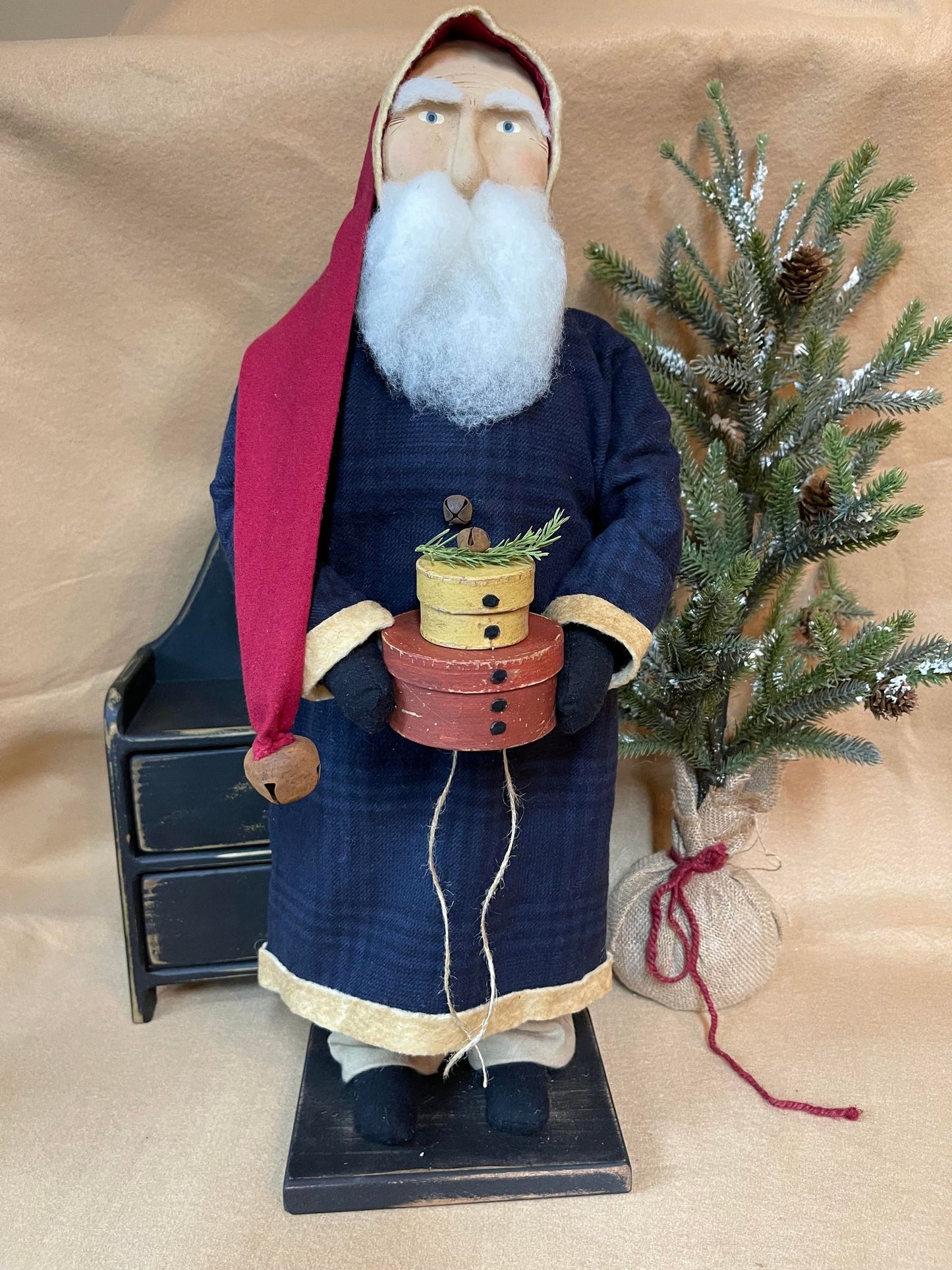 A Belsnickel figurine with a blue jacket, red hat, and white beard, holding two pantry boxes, standing next to a Christmas tree and a decorative bench.