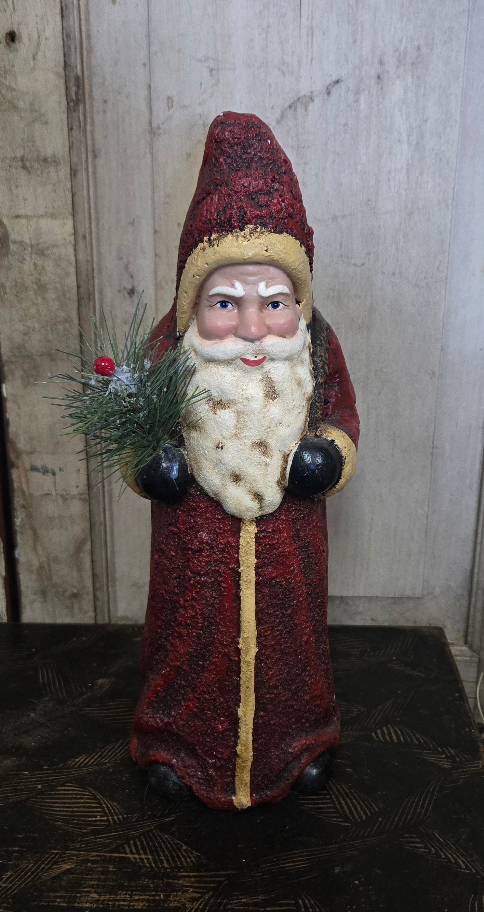 Decorative Santa Claus figure with a red beard and hat, holding a pine branch, against a wooden background.