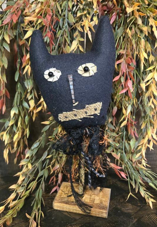 Textile art piece resembling a cat with a skull face on a wooden stand, surrounded by greenery.
