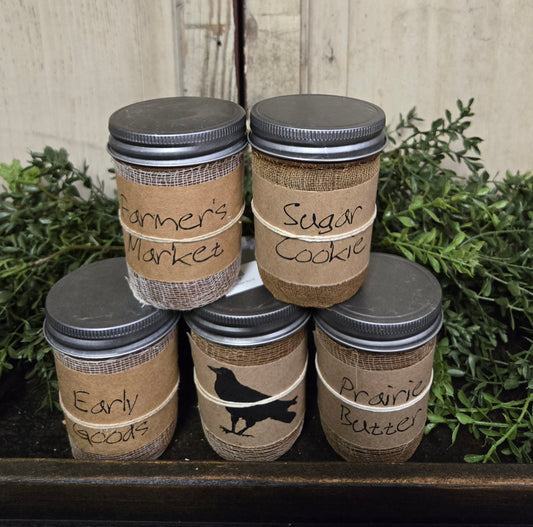 A set of Black Crow jar candles with various labels, placed on a surface with greenery in the background.