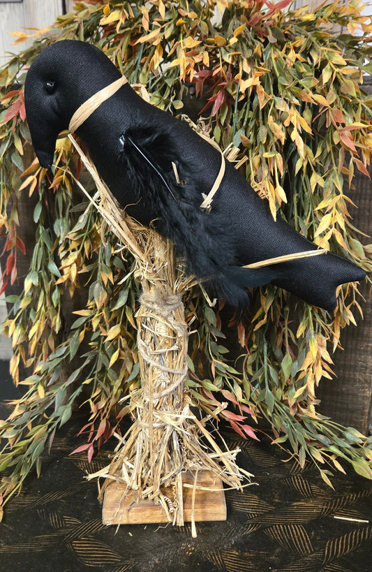 Decorative black cat figure with straw body on a wooden base, surrounded by greenery.