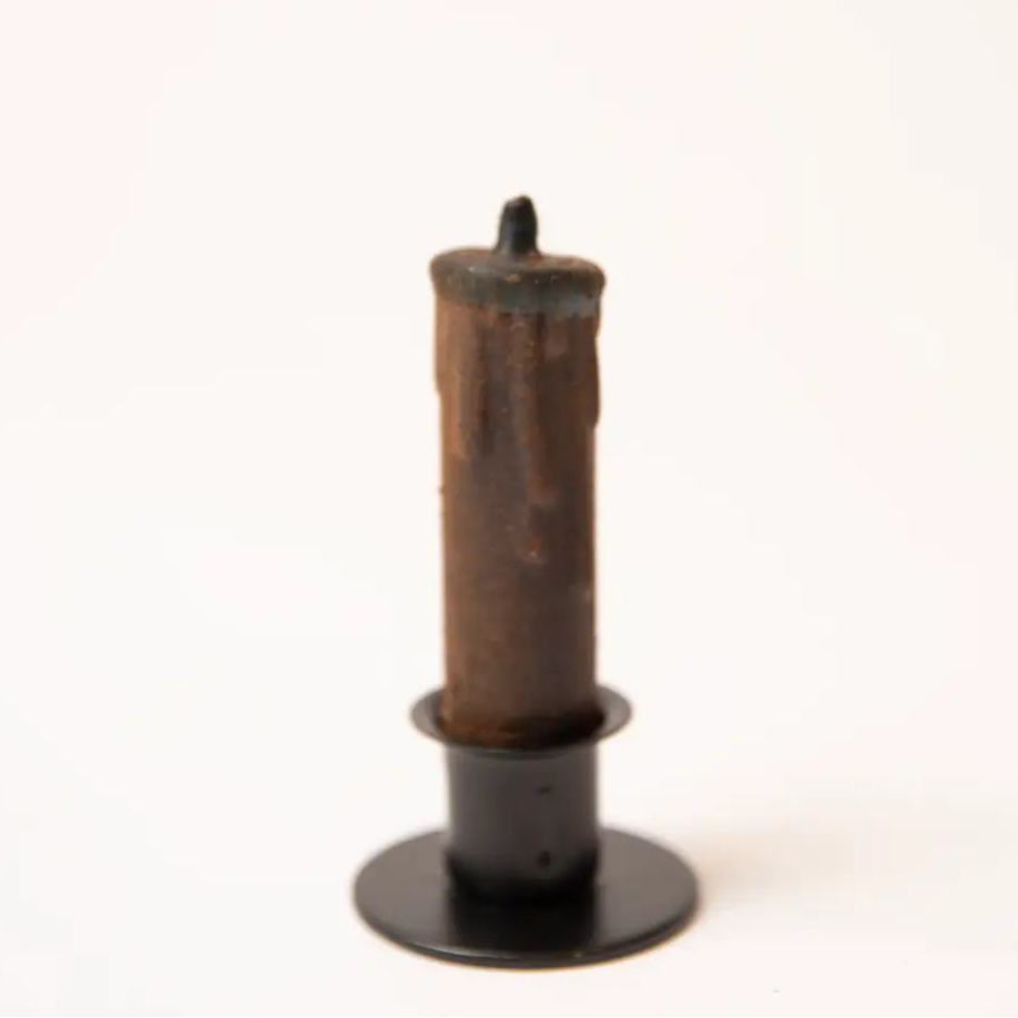 A single old-style taper candle with a heavily weathered, dark brown and black wax surface, standing in a black metal holder against a plain white background.