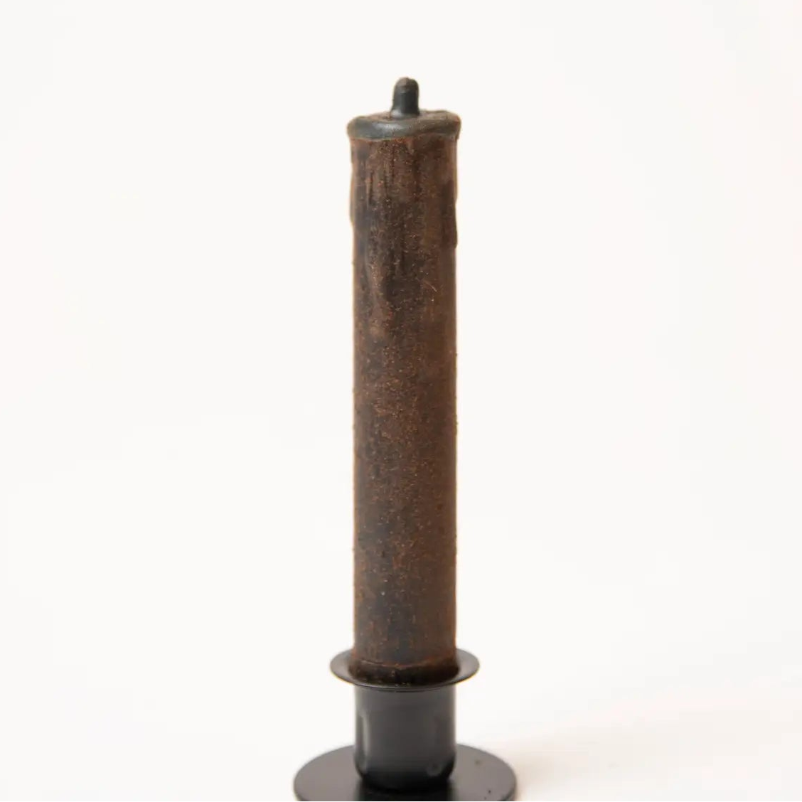 A single dark brown candle with a textured, rustic finish, standing upright in a black metal holder against a plain white background.