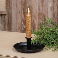A twisted taper candle with a lit flame stands in a black holder on a round black tray, set on a tabletop with a rustic wooden background and greenery.