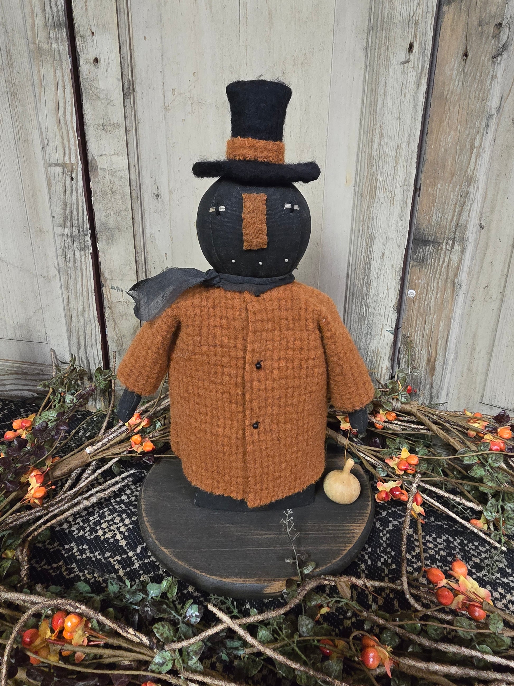 Pumpkin head doll with orange coat on a black riser among bittersweet