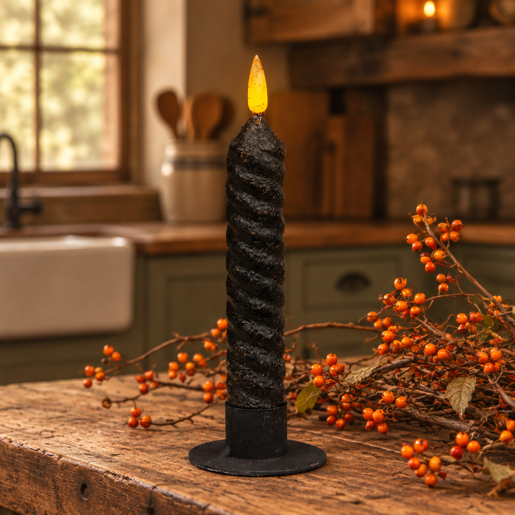 A black twisted spiral taper candle with a glowing yellow flame sits on a rustic wooden table in a warm, softly blurred kitchen setting, surrounded by orange berries.
