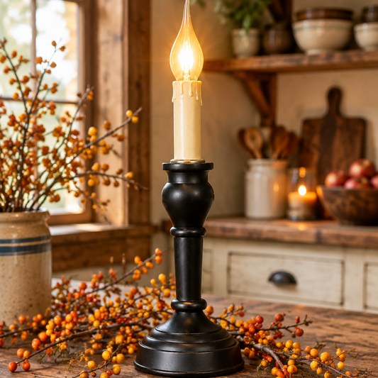 A black metal candlestick holder with a lit cream-colored candle sits on a rustic wooden table, surrounded by autumn berry branches in a cozy, warmly lit kitchen setting.
