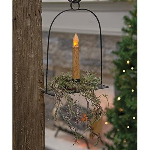 A black hanging candle pan with a candle lit, placed against a backdrop of a Christmas tree and holiday decorations.