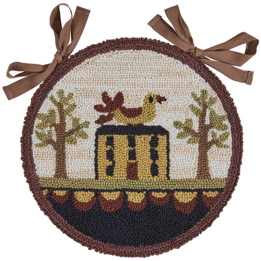 A round chair pad with a 'Bless Our Home' design, featuring an artistic representation of a house, trees, and a chicken, in a hooked rug style.