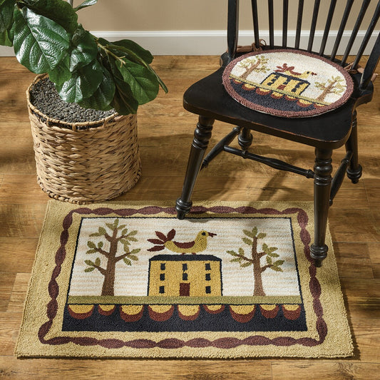 A rectangular hooked rug with a house and trees design, placed on the floor next to a chair and a basket.