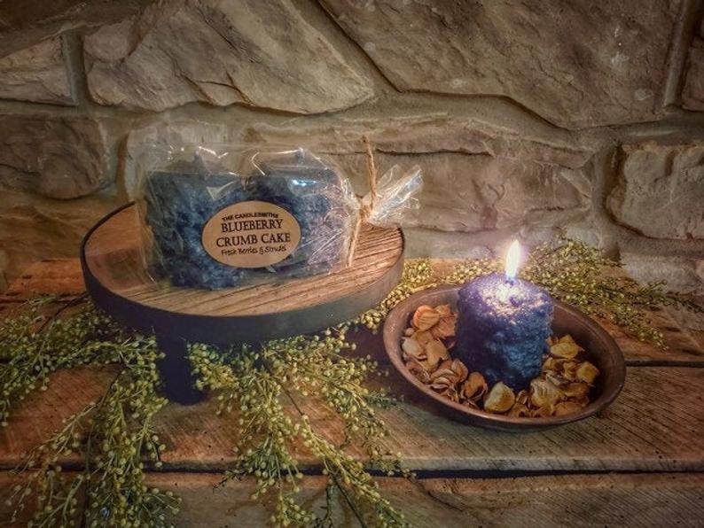 Blue candle burning in a wooden bowl with potpourri next to a riser with two candles in packaging