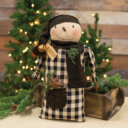 A stuffed snowman with a plaid pattern, wearing a dark brown scarf and hat, standing in front of a wooden structure with artificial pine sprigs and a brown paper tag that reads 'Buster'.