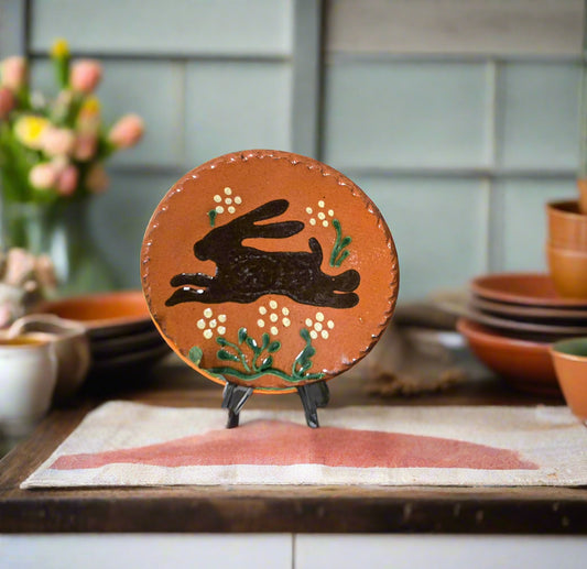 Small round redware plate with brown bunny and flowers sitting among greenery