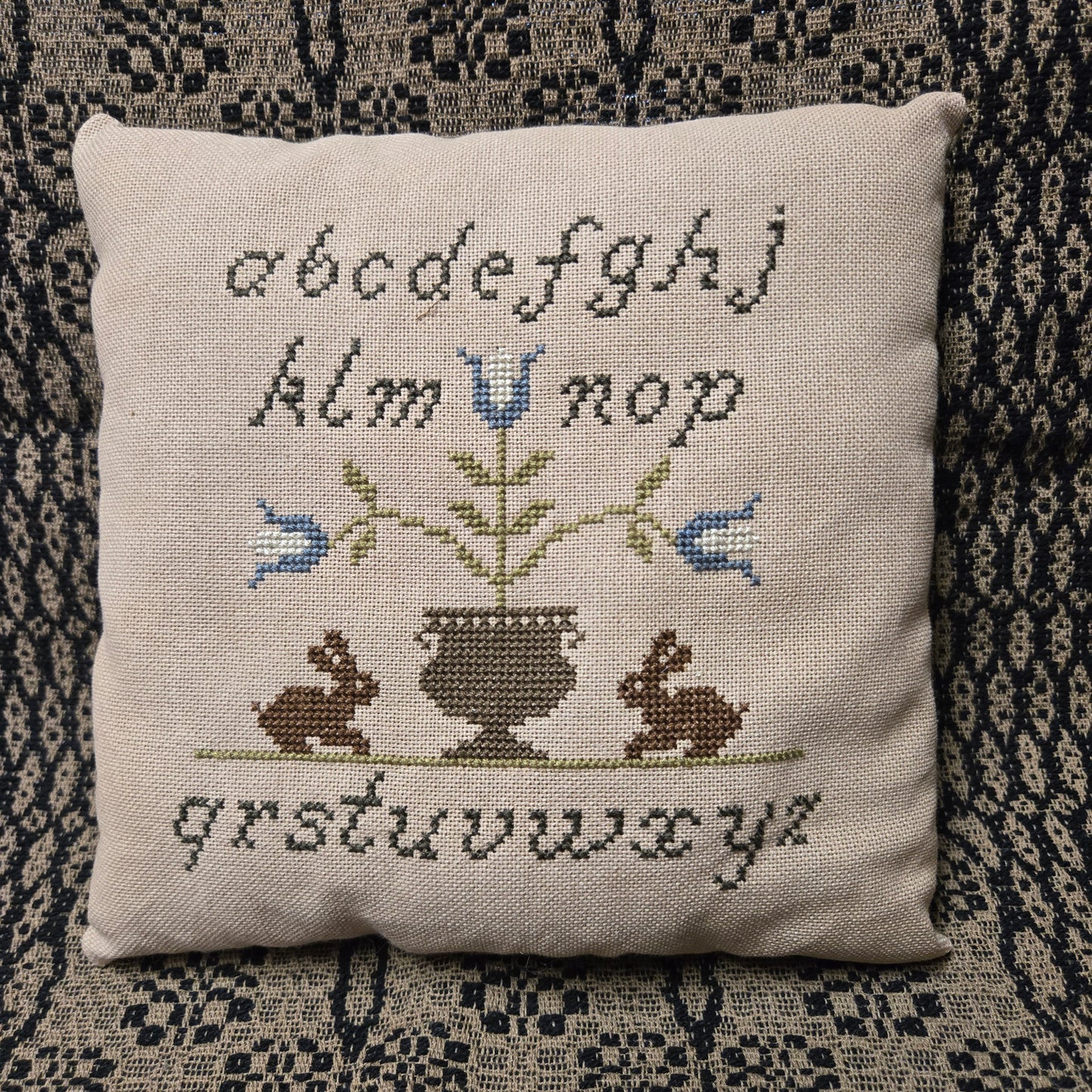 Decorative pillow with cross-stitched designs and text on a textured surface